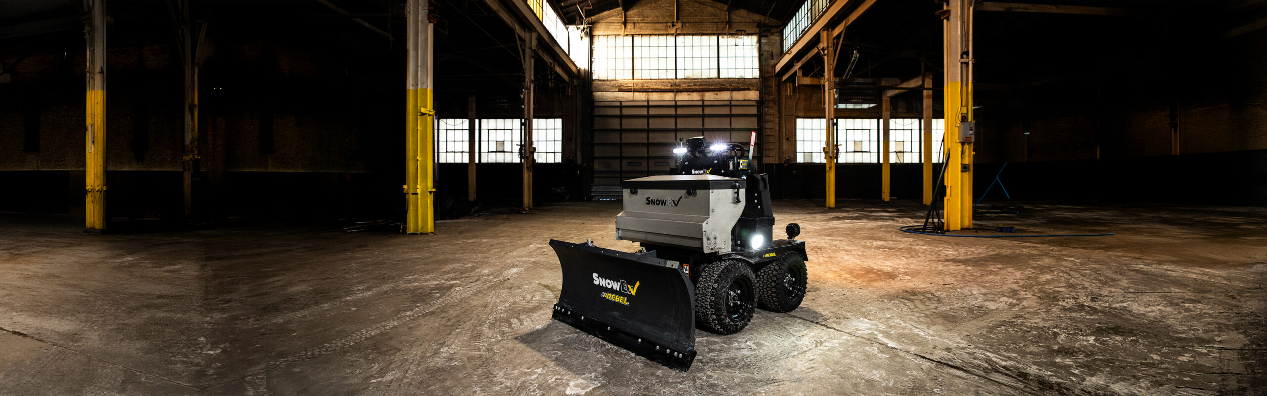 A compact, autonomous construction vehicle with a plow attachment is parked in a large, empty industrial warehouse with yellow pillars and high windows.