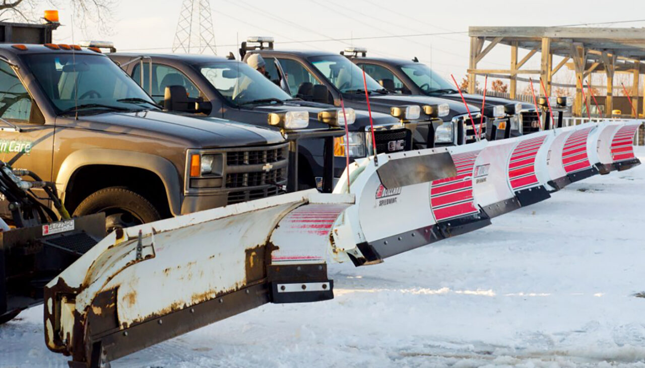A row of black pickup trucks with large snow plows attached to their fronts are parked on a snow-covered lot under a clear sky.