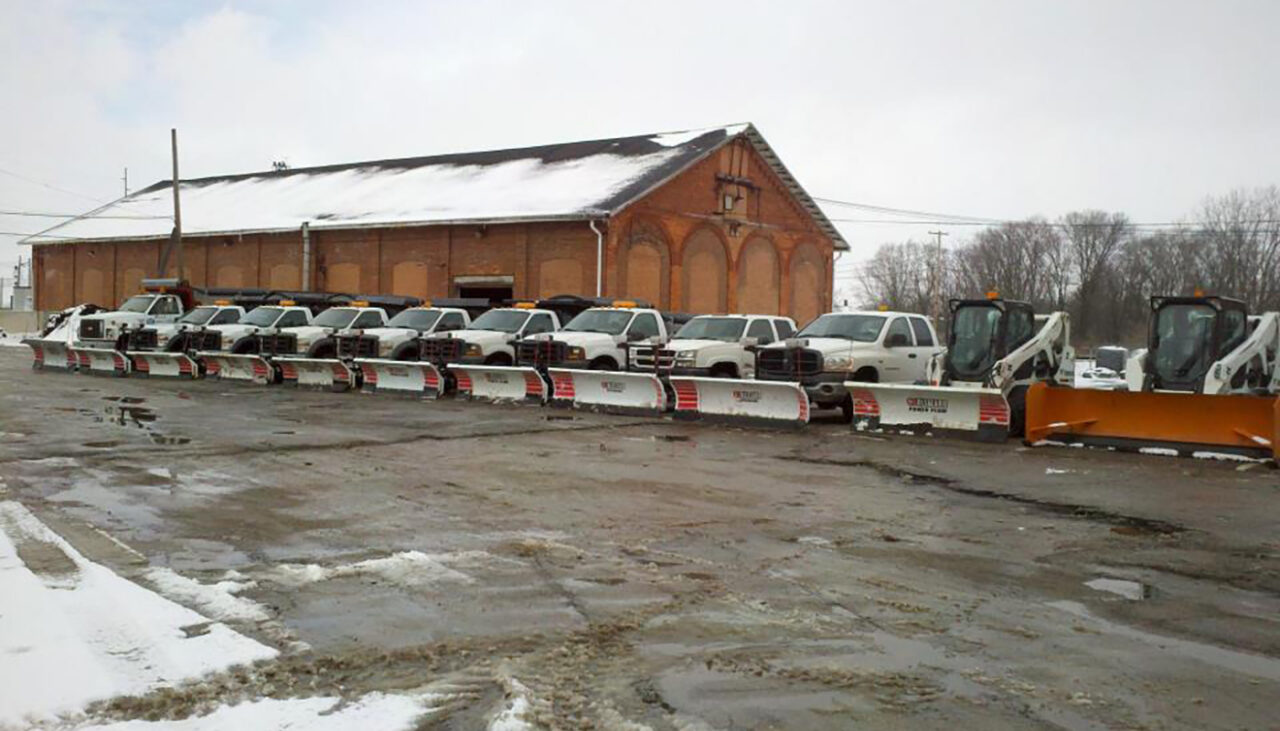 A row of snow plow trucks and skid steers with plows attached are parked on a wet, partially snow-covered lot in front of a large brick building on a cloudy winter day.